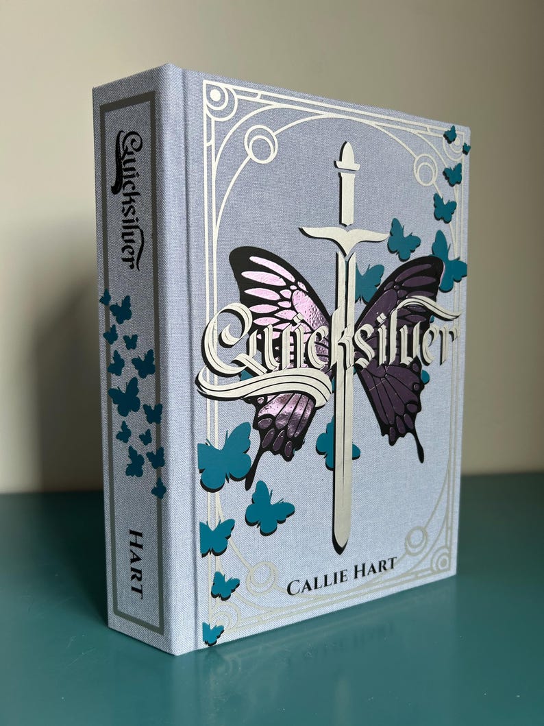 May include: A hardback book titled "Quicksilver" by Callie Hart. The cover is light blue with a silver sword and butterfly design. Teal butterflies and the author's name are also visible on the cover.