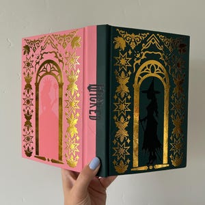 Puede incluir: Un libro decorativo con cubiertas rosa y verde. La cubierta rosa presenta un diseño de arco dorado con una silueta, mientras que la cubierta verde tiene una silueta de bruja. La columna vertebral dice "WICKED".