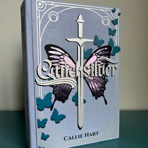 May include: A hardback book titled "Quicksilver" by Callie Hart. The cover is light blue with a design featuring a sword and butterfly wings. Teal butterflies are scattered around the cover.