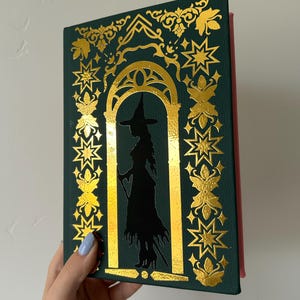May include: A dark green book with gold foil accents and a silhouette of a witch. The cover features an arched doorway design with star and floral patterns. The book's spine is red.