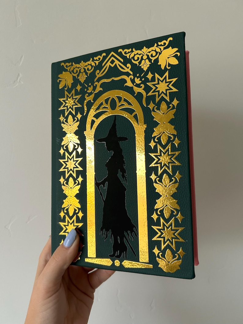 May include: A dark green book with gold foil accents and a silhouette of a witch. The cover features an arched doorway design with star and floral patterns. The book's spine is red.