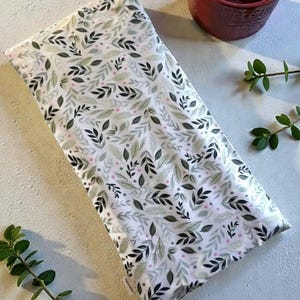 May include: A rectangular heating pad featuring a white fabric cover with a repeating pattern of dark green, sage green, and light gray leaves interspersed with small pink dots, accompanied by a dark red ceramic mug and green sprigs.