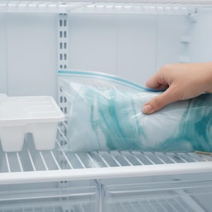 Puede incluir: Interior de un refrigerador con una cubitera blanca y una bolsa de pl&aacute;stico transparente que contiene un art&iacute;culo azul y blanco. Una mano se acerca a la bolsa. El refrigerador tiene estantes de alambre.