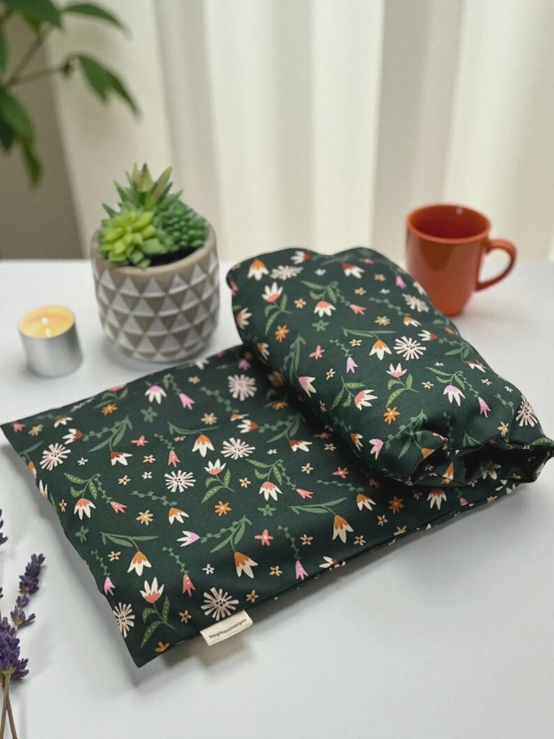 Puede incluir: Dos almohadillas t&eacute;rmicas con estampado floral verde oscuro. Las almohadillas tienen un patr&oacute;n de flores blancas, rosas y naranjas con hojas verdes. Una peque&ntilde;a planta suculenta en una maceta gris, una vela y una taza est&aacute;n en el fondo.