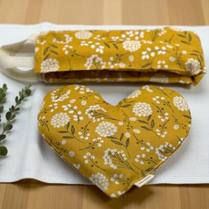May include: Mustard yellow heart-shaped pillow and matching rectangular eye mask. Both items feature a white floral pattern with dark green stems and leaves. The eye mask has a cream-colored strap. The items are placed on a white surface.