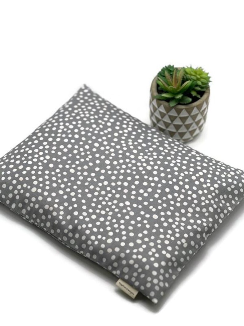 K&ouml;nnte beinhalten: Ein graues, quadratisches Kissen mit wei&szlig;em Tupfenmuster. Daneben steht ein kleiner Topf mit geometrischem Muster, der eine Sukkulentenpflanze enth&auml;lt. Das Kissen hat ein kleines Etikett.