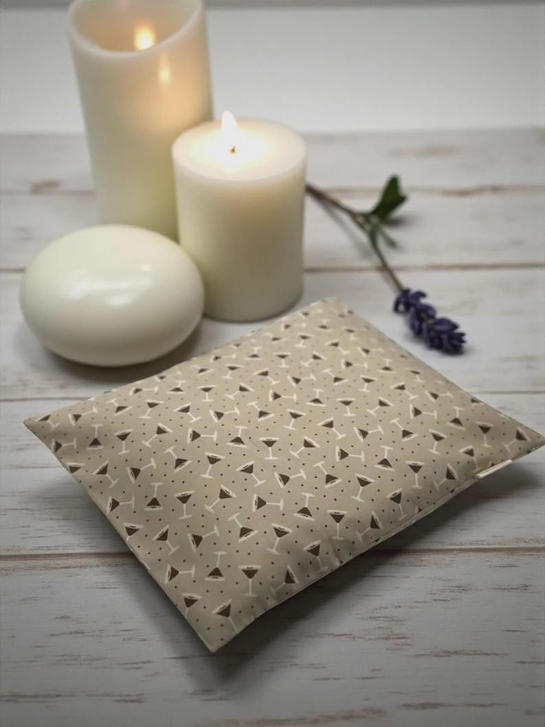 K&ouml;nnte beinhalten: Ein beiges Kissen mit Martini-Gl&auml;ser-Muster, brennenden Kerzen und einer wei&szlig;en Seife auf einer Holzoberfl&auml;che. Das Kissen hat ein sich wiederholendes Muster aus wei&szlig;en Martini-Gl&auml;sern mit braunen Details. Zwei brennende Kerzen und eine wei&szlig;e Seife sind im Hintergrund.