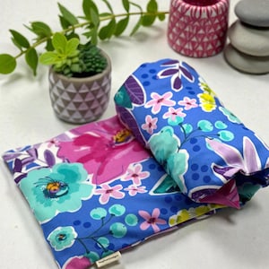 Puede incluir: Dos art&iacute;culos de tela con estampado floral, uno doblado y otro plano, con un fondo azul vibrante y flores rosas, moradas y turquesas. El art&iacute;culo doblado est&aacute; enrollado. Una peque&ntilde;a planta en maceta y objetos decorativos est&aacute;n en el fondo.