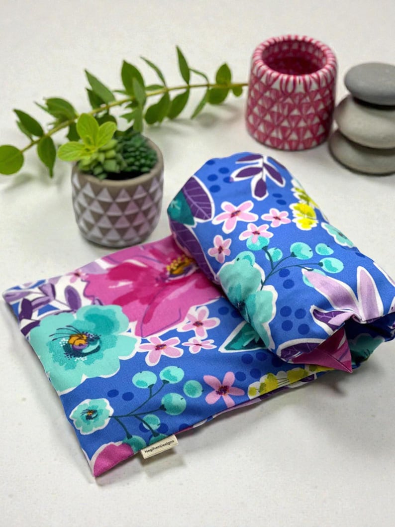Puede incluir: Dos art&iacute;culos de tela con estampado floral, uno doblado y otro plano, con un fondo azul vibrante y flores rosas, moradas y turquesas. El art&iacute;culo doblado est&aacute; enrollado. Una peque&ntilde;a planta en maceta y objetos decorativos est&aacute;n en el fondo.
