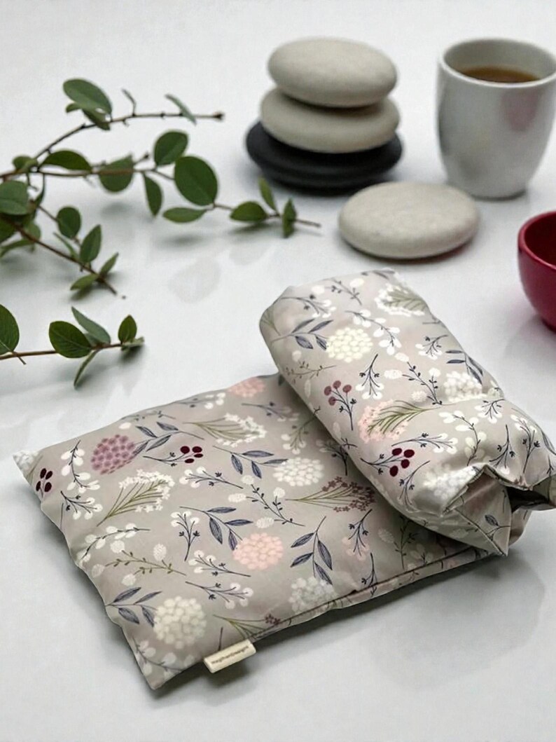 Puede incluir: Se muestran dos almohadillas t&eacute;rmicas grises con estampado floral, una rectangular y otra cil&iacute;ndrica. Las almohadillas presentan un patr&oacute;n de flores blancas, rosas y burdeos con hojas verdes. Las almohadillas est&aacute;n sobre una superficie blanca.
