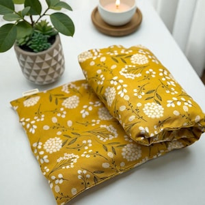 Puede incluir: Dos almohadas rectangulares de color amarillo mostaza con un estampado floral blanco y tallos verde oscuro. Una peque&ntilde;a planta en maceta y una vela encendida en una taza blanca sobre un posavasos de madera tambi&eacute;n son visibles sobre una superficie blanca.