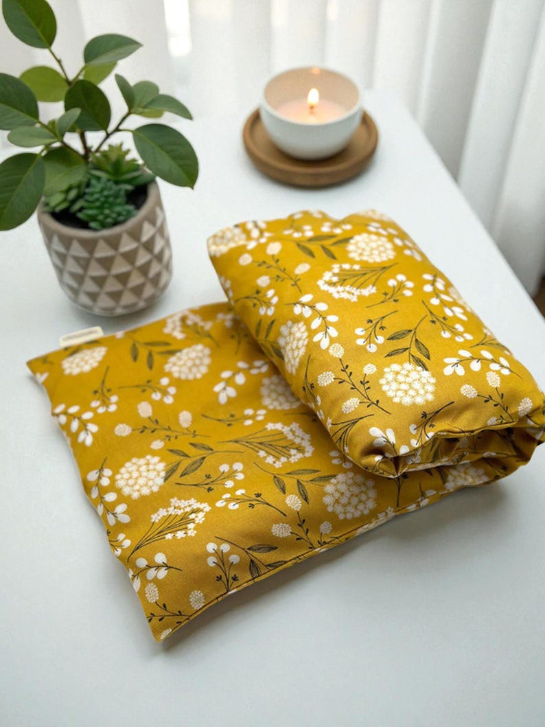 Puede incluir: Dos almohadas rectangulares de color amarillo mostaza con un estampado floral blanco y tallos verde oscuro. Una peque&ntilde;a planta en maceta y una vela encendida en una taza blanca sobre un posavasos de madera tambi&eacute;n son visibles sobre una superficie blanca.