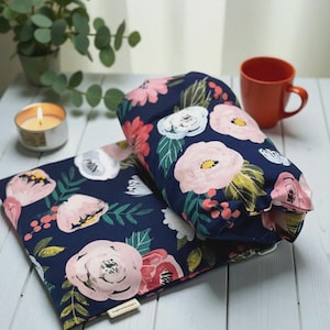 Puede incluir: Dos almohadas con estampado floral azul marino con flores rosas y blancas y hojas verdes. Una taza naranja, una vela encendida y una peque&ntilde;a planta en maceta est&aacute;n en el fondo. Las almohadas est&aacute;n sobre una superficie de madera blanca.