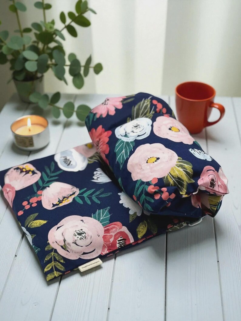 Puede incluir: Dos almohadas con estampado floral azul marino con flores rosas y blancas y hojas verdes. Una taza naranja, una vela encendida y una peque&ntilde;a planta en maceta est&aacute;n en el fondo. Las almohadas est&aacute;n sobre una superficie de madera blanca.