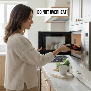Puede incluir: Una persona colocando comida en un horno microondas negro en una cocina. El microondas est&aacute; empotrado en gabinetes blancos. El texto encima del microondas dice "DO NOT OVERHEAT". Una taza y un libro est&aacute;n en la encimera.