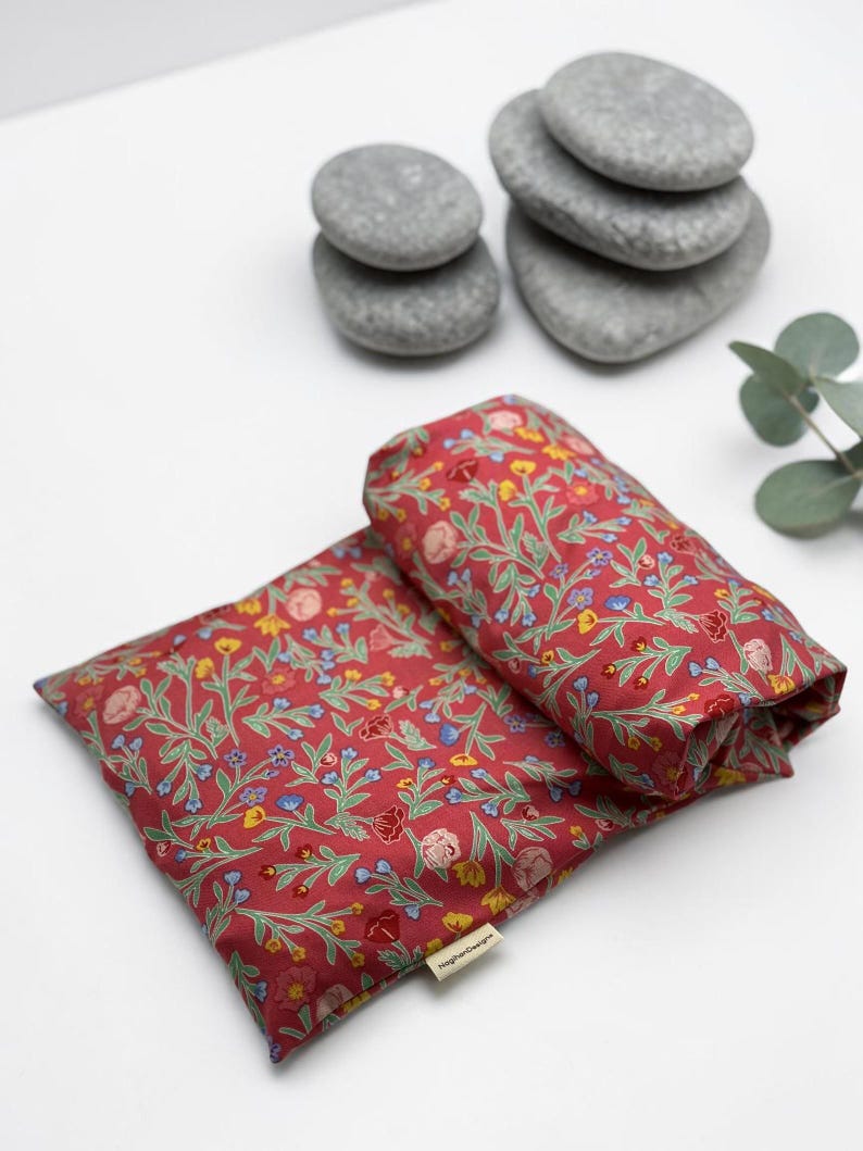 Puede incluir: Dos almohadas rojas con estampado floral, una doblada, con un patr&oacute;n de flores y hojas amarillas, azules y rosas. Piedras grises y una ramita de vegetaci&oacute;n en el fondo. Las almohadas tienen una etiqueta que dice "Higher Designs".