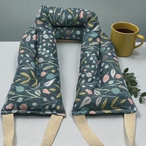May include: A dark teal neck wrap with a floral pattern in pink, yellow, and white. The wrap has cream-colored straps and is next to a yellow mug of tea. A sprig of greenery is on the table.