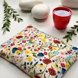 K&ouml;nnte beinhalten: Ein rechteckiges Kissen mit floralem Muster, cremefarbenem Hintergrund und roten, gelben, blauen und rosa Blumen. Eine kleine rote Tasse mit dunkler Fl&uuml;ssigkeit steht in der N&auml;he von wei&szlig;en Steinen und gr&uuml;nen Zweigen. Das Kissen scheint gef&uuml;llt zu sein.