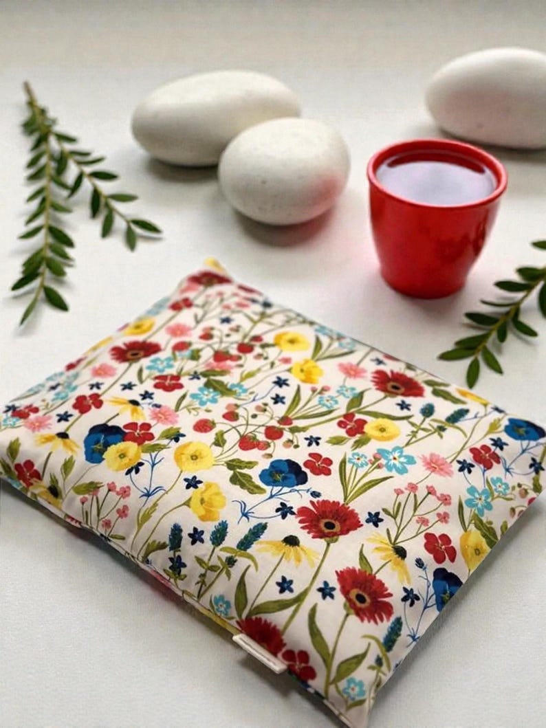 K&ouml;nnte beinhalten: Ein rechteckiges Kissen mit floralem Muster, cremefarbenem Hintergrund und roten, gelben, blauen und rosa Blumen. Eine kleine rote Tasse mit dunkler Fl&uuml;ssigkeit steht in der N&auml;he von wei&szlig;en Steinen und gr&uuml;nen Zweigen. Das Kissen scheint gef&uuml;llt zu sein.