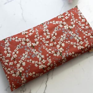 May include: A rectangular pillow featuring a rust-colored fabric base with a repeating pattern of white cherry blossom branches and small light blue floral accents. The pillow has a small fabric tag on the side edge and a smooth texture.