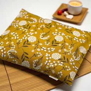 K&ouml;nnte beinhalten: Senfgelbes Kissen mit wei&szlig;em Blumenmuster. Das Kissen hat ein sich wiederholendes Design aus wei&szlig;en Blumen und dunkelgr&uuml;nen Bl&auml;ttern. Das Kissen ist rechteckig und scheint aus Stoff zu sein. Das Kissen liegt auf einer Bambusmatte.