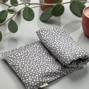 Puede incluir: Dos almohadas de tela gris con lunares blancos. Una almohada es rectangular y plana, mientras que la otra est&aacute; enrollada. Las almohadas est&aacute;n sobre una superficie gris claro, con hojas verdes y dos tazas de color terracota en el fondo.