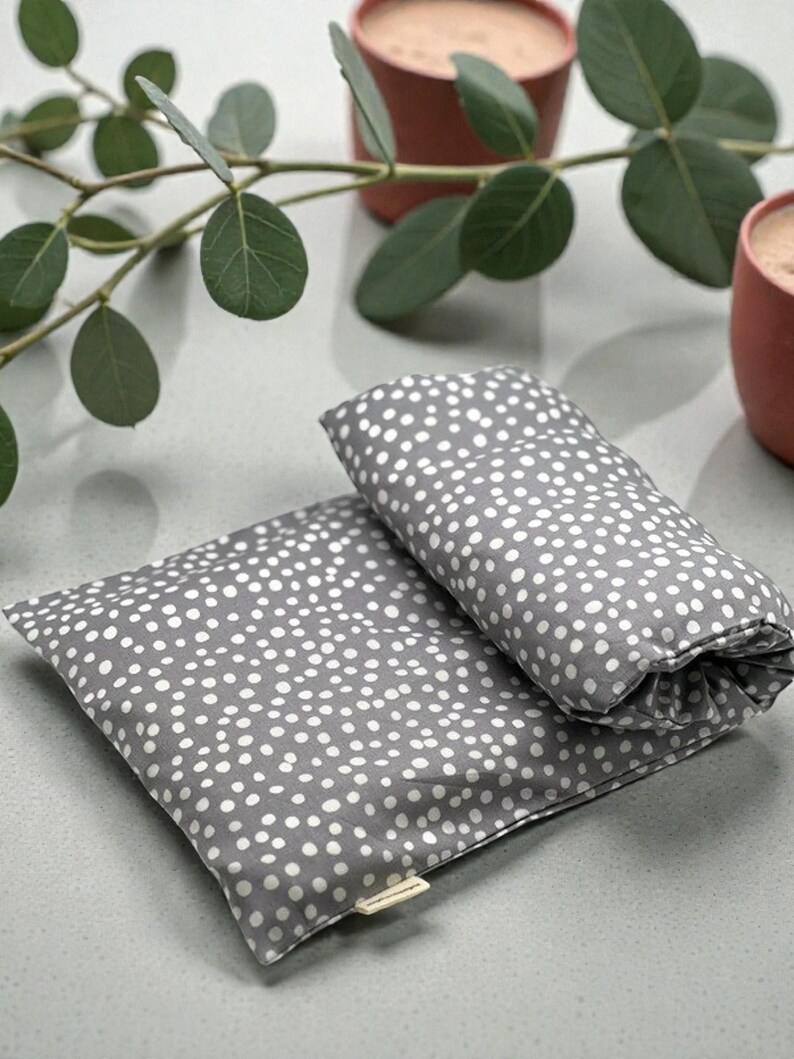 Puede incluir: Dos almohadas de tela gris con lunares blancos. Una almohada es rectangular y plana, mientras que la otra est&aacute; enrollada. Las almohadas est&aacute;n sobre una superficie gris claro, con hojas verdes y dos tazas de color terracota en el fondo.