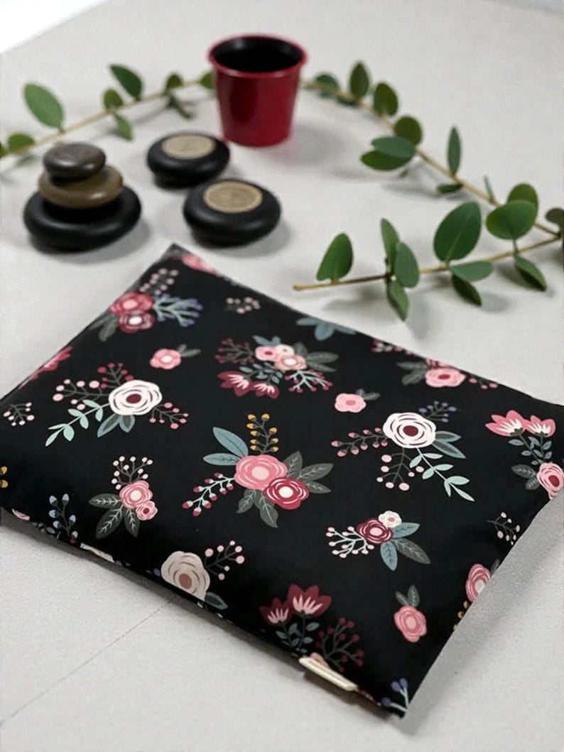 K&ouml;nnte beinhalten: Ein schwarzes rechteckiges Kissen mit einem Blumenmuster aus rosa und wei&szlig;en Rosen, gr&uuml;nen Bl&auml;ttern und Beeren. Das Bild zeigt auch einen kleinen roten Topf, gestapelte Steine und Zweige mit Gr&uuml;n.