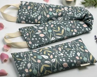May include: Three rectangular, dark teal fabric aromatherapy pillows with a floral pattern of white, pink, yellow, and teal. Each pillow has a cream-colored fabric handle. A lit candle and rose petals are in the background.