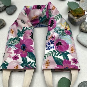 May include: A long, rectangular fabric neck wrap featuring a floral pattern with shades of pink, magenta, and dark green on a white background. The wrap has two beige fabric handles attached to the ends, allowing for easy handling and positioning.