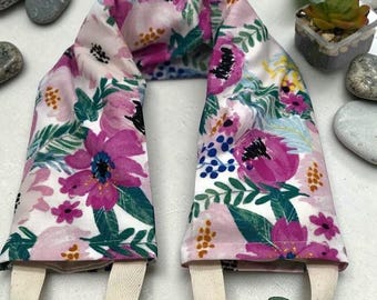 Puede incluir: Un envoltorio para el cuello con estampado floral sobre fondo blanco, con grandes flores rosas y moradas, hojas verdes y detalles amarillos. Tiene lazos de tela color crema en cada extremo. El producto está diseñado para la comodidad y la relajación.