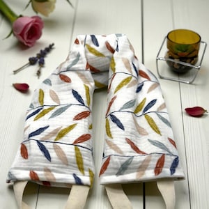 May include: A white fabric neck wrap with a colorful leaf pattern in shades of blue, yellow, and red. The wrap has two beige straps and is designed for comfort and relaxation. A candle holder and flowers are in the background.