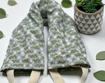 May include: A light blue fabric heating pad featuring a repeating pattern of small white flowers and olive green leaves. The rectangular item has two beige cotton webbing handles attached at the bottom, allowing for easy handling and positioning.