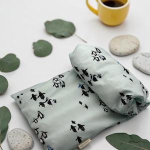Puede incluir: Un coj&iacute;n azul claro y tela enrollada con un dise&ntilde;o de ping&uuml;inos. Una taza amarilla con caf&eacute;, hojas de eucalipto y peque&ntilde;as piedras est&aacute;n dispuestas sobre una superficie blanca.