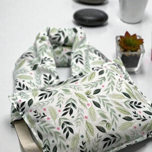 May include: A white and green leaf-patterned neck wrap and square pillow. The fabric features a repeating design of green leaves and pink accents. A cream-colored strap is attached to the pillow. The items are likely for relaxation or therapeutic use.