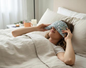 May include: A person lying in bed wearing a blue and white floral patterned eye mask. The mask is held in place, covering the eyes. The bedding is a neutral color, and the scene suggests relaxation and sleep.