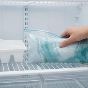 K&ouml;nnte beinhalten: Innenraum eines K&uuml;hlschranks mit einem wei&szlig;en Eisw&uuml;rfelbeh&auml;lter und einem transparenten Plastikbeutel mit einem blau-wei&szlig;en Gegenstand. Eine Hand greift nach dem Beutel. Der K&uuml;hlschrank hat Drahtregale.
