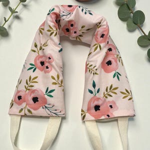 May include: A U-shaped fabric neck wrap featuring a light pink background with a floral pattern of soft pink flowers, dark green leaves, and thin stems. The wrap includes two off-white fabric loops attached to the bottom ends for handling.