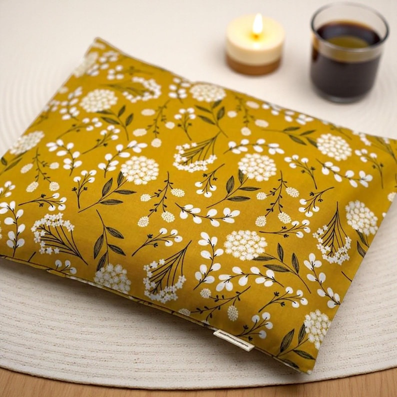K&ouml;nnte beinhalten: Senfgelbes rechteckiges Kissen mit wei&szlig;em Blumenmuster. Das Kissen ist mit wei&szlig;en Bl&uuml;ten und dunkelgr&uuml;nen Bl&auml;ttern verziert. Eine brennende Kerze und ein Glas dunkler Fl&uuml;ssigkeit befinden sich im Hintergrund.