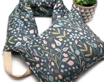 May include: A dark teal floral patterned neck wrap and square pillow set. The fabric features a botanical design with pink, yellow, and blue accents. The neck wrap has a beige strap. A small potted succulent is in the background.