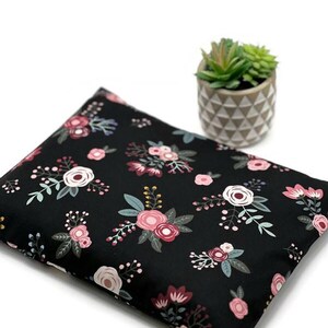 K&ouml;nnte beinhalten: Ein schwarzes Kissen mit einem Blumenmuster aus rosa, wei&szlig;en und bordeauxfarbenen Blumen mit gr&uuml;nen Bl&auml;ttern. Das Kissen ist rechteckig und liegt auf einer wei&szlig;en Oberfl&auml;che. Eine kleine Topfpflanze mit Sukkulenten befindet sich im Hintergrund.
