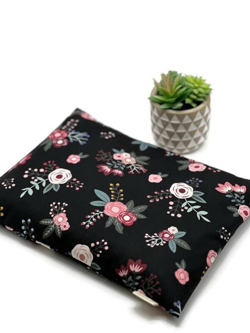 K&ouml;nnte beinhalten: Ein schwarzes Kissen mit einem Blumenmuster aus rosa, wei&szlig;en und bordeauxfarbenen Blumen mit gr&uuml;nen Bl&auml;ttern. Das Kissen ist rechteckig und liegt auf einer wei&szlig;en Oberfl&auml;che. Eine kleine Topfpflanze mit Sukkulenten befindet sich im Hintergrund.