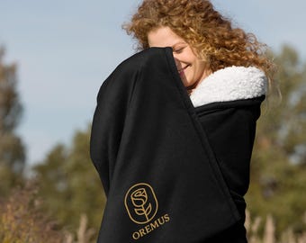 May include: A black hooded blanket with a white sherpa lining. The blanket has a gold rose logo and the word "OREMUS". The person in the image is smiling and wrapped in the blanket.
