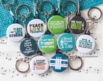 Puede incluir: Un conjunto de 12 llaveros coloridos con diferentes frases sobre el tema de la trompeta. Los llaveros están sobre una hoja de música con un fondo azul.