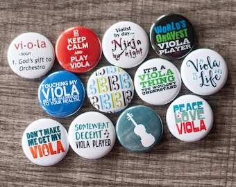 May include: A collection of 11 round buttons with various viola-themed designs. The buttons are white with black, red, green, blue, and yellow text and images. Some of the text includes "Keep Calm and Play Viola", "World's Okayest Viola Player", "It's a Viola Thing", and "Peace Love Viola".