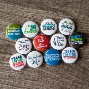 Puede incluir: Una colección de 12 botones redondos de colores con diferentes frases sobre el tema del saxofón. Los botones presentan frases como "Plays Well With Others", "World's Okayest Saxophone Player", "Keep Calm and Play Saxophone", y "Saxophone is Life".