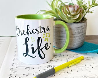 May include: White ceramic mug with a green rim and handle. The mug has black text that reads "Orchestra is Life" with musical notes in yellow. The mug is sitting on a sheet of music with a pencil.