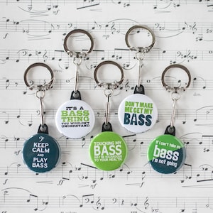 May include: Five keychains with round, colorful buttons featuring bass-themed puns. The buttons are green, white, and black with white text. The keychains are silver with black clasps.