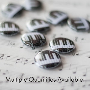 May include: Multiple piano key design buttons on a sheet of music. The buttons are black and white with a silver backing. The text "www.charmandcassie.com" is visible on one of the buttons.