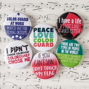 May include: Nine colorful round buttons with different slogans related to color guard. The slogans include "Color Guard at Work", "Peace Love Color Guard", "I Didn't Choose Colorguard Colorguard Chose Me", "If You Can't Spin Don't Touch My Flag", "I Have a Life I Just Like Guard Better", and "There Are Two Types of People in the World: In Color Guard, and Those Who Want to Be."